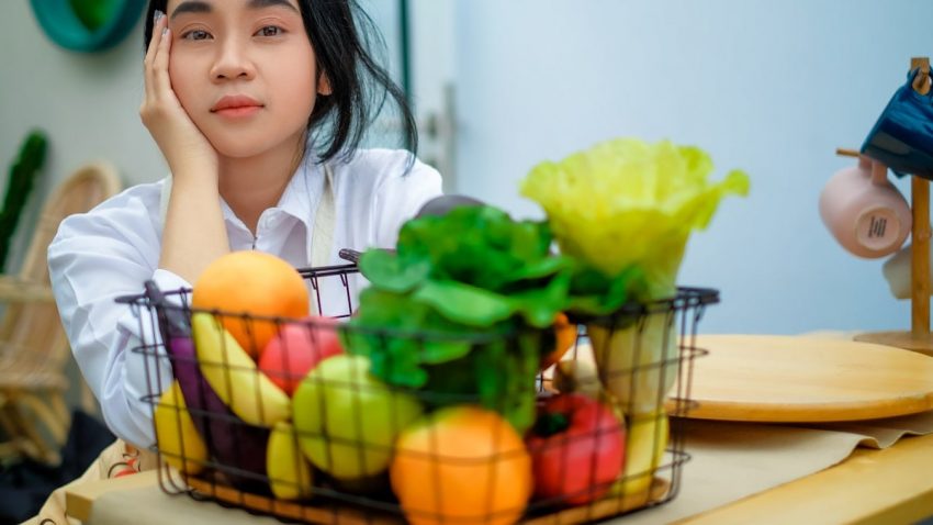 a girl sitting at a table with a basket of fruit and vegetables